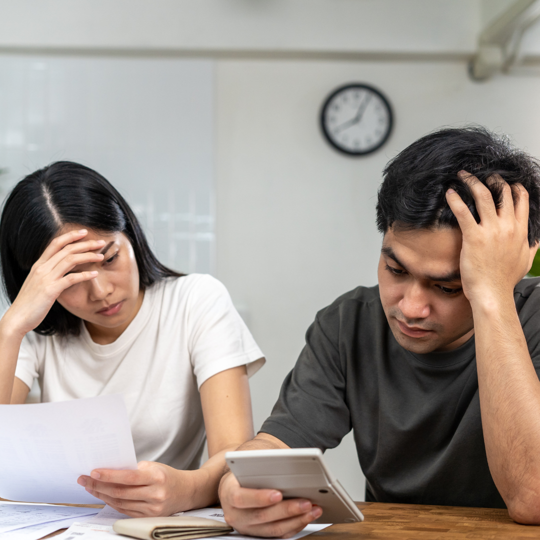 Two people sit at a table looking stressed while looking at bills and a calculator.