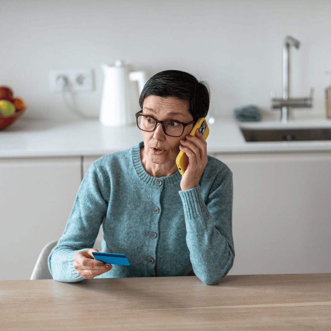 A person wearing glasses and a blue cardigan talks on a yellow phone while holding a blue card at a wooden table.