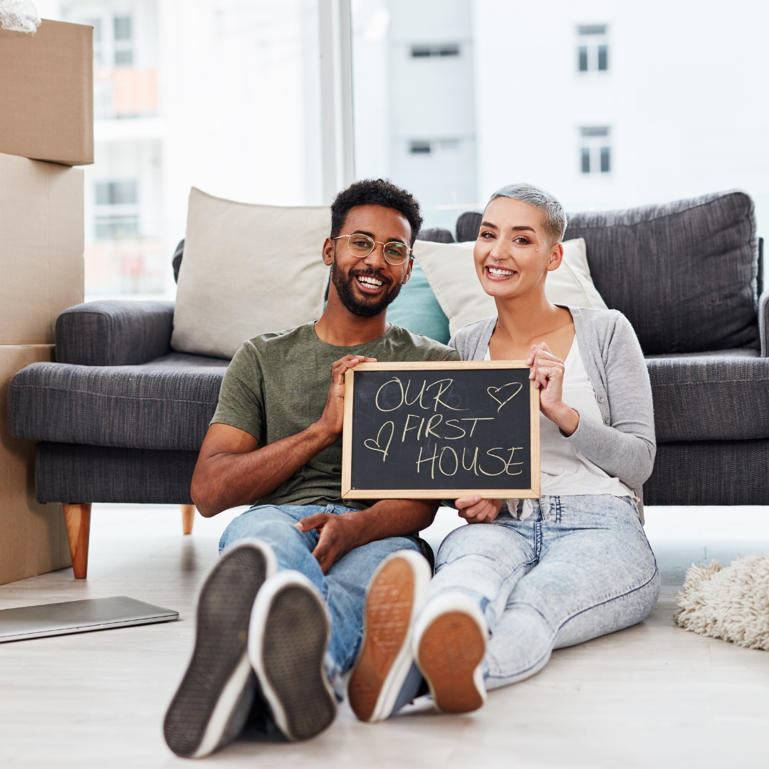 People sitting on the floor in front of a couch, holding a sign that reads "Our First House".