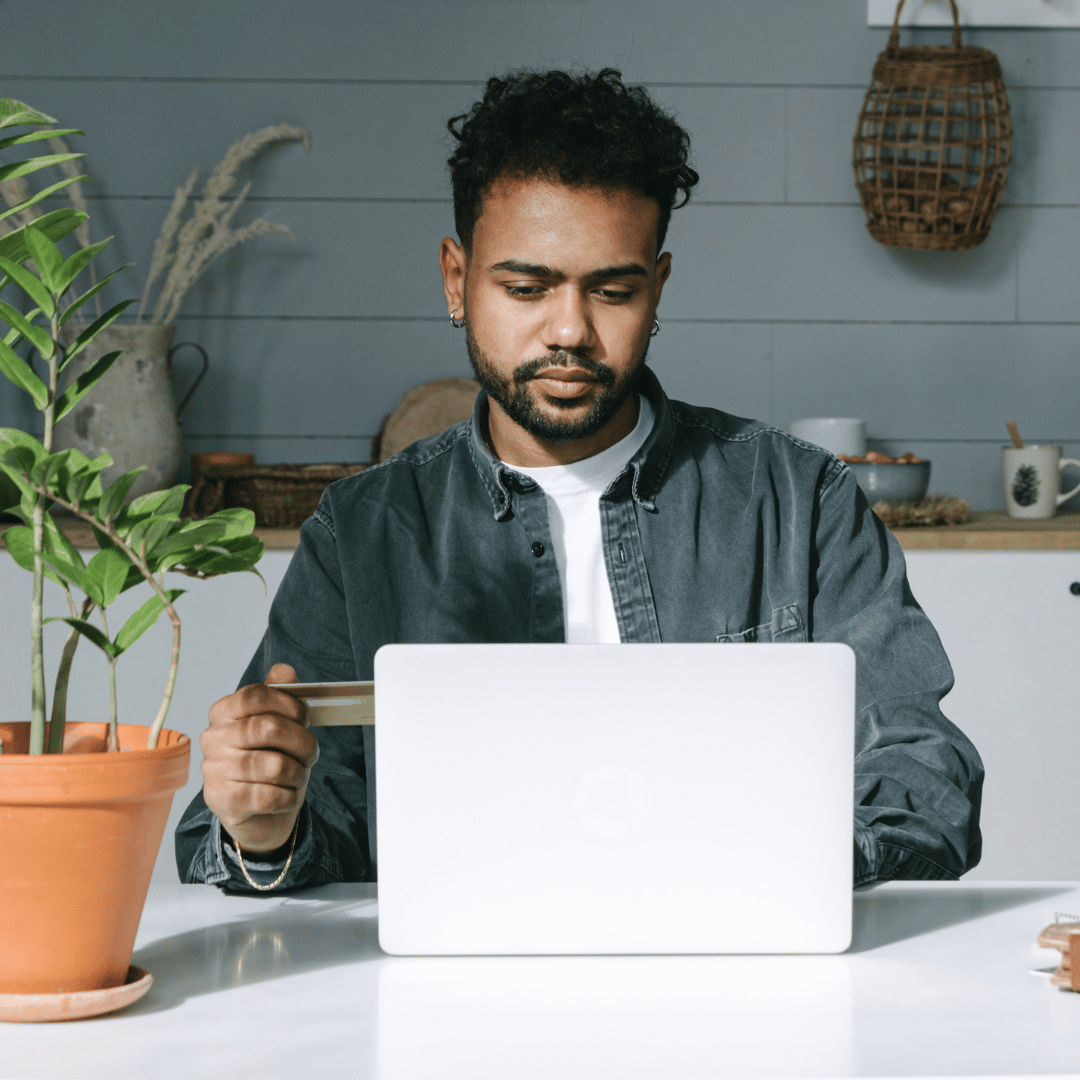 A person sits at a table with a laptop and holds a credit card, appearing to make an online purchase.