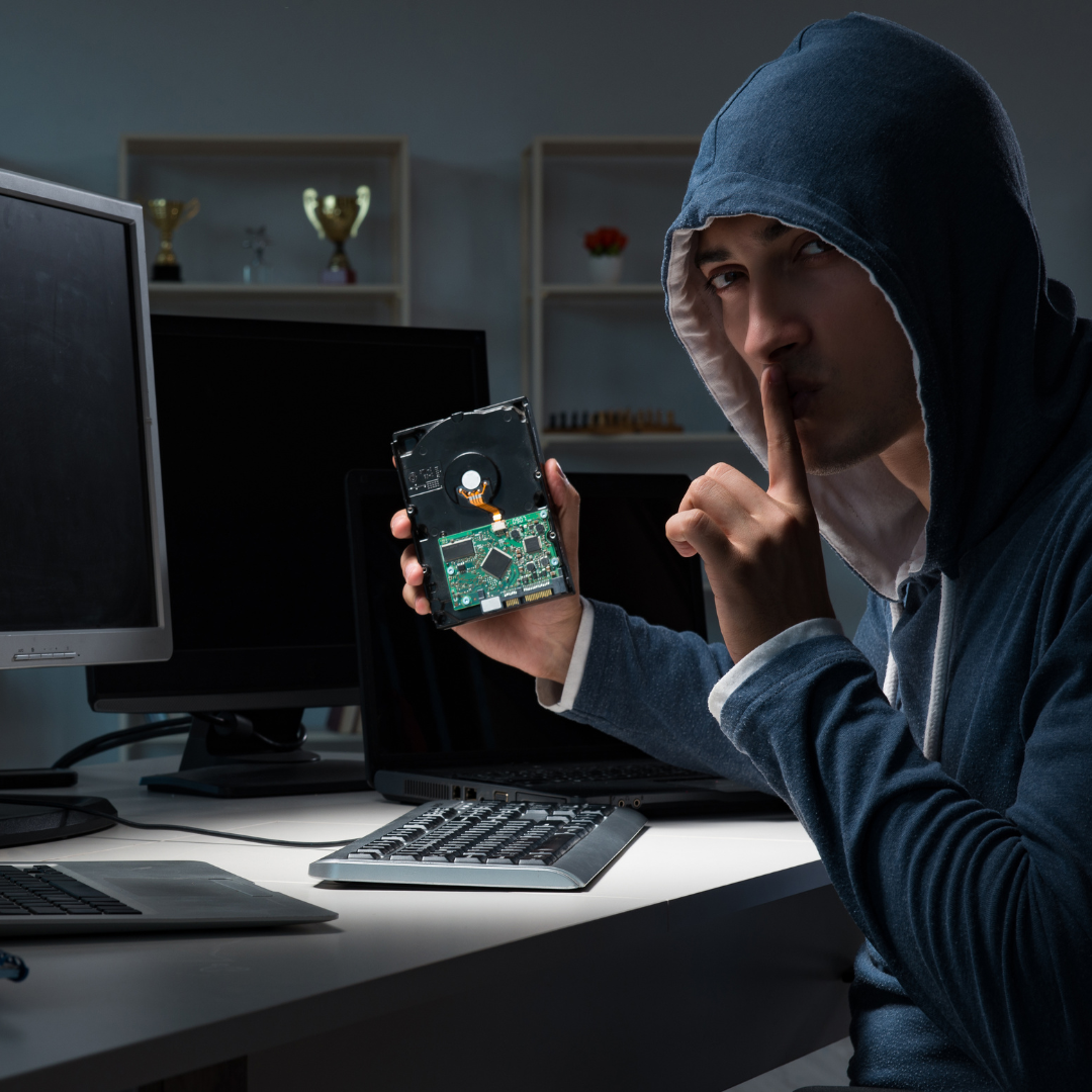A person in a hoodie holds up a hard drive, placing a finger to their lips in a gesture for silence, in front of computer screens.
