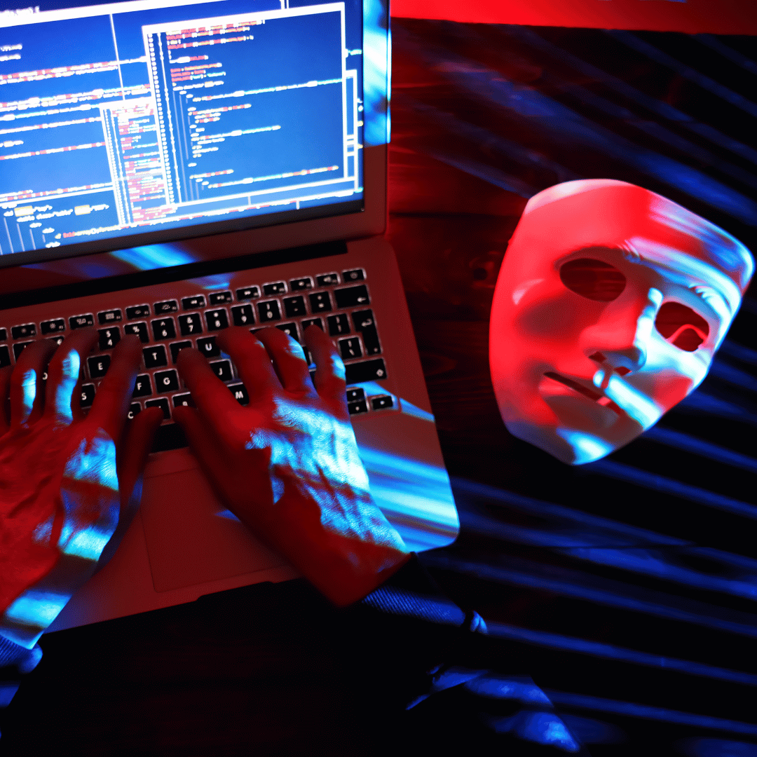 A white mask rests on a dark surface next to a laptop with code on the screen, illuminated by red and blue lights.