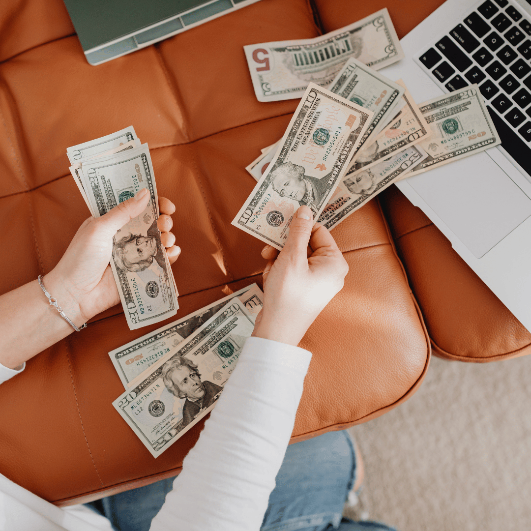 Hands holding several US dollar bills while other cash is scattered on an orange couch, with a laptop keyboard and a wallet in the background.