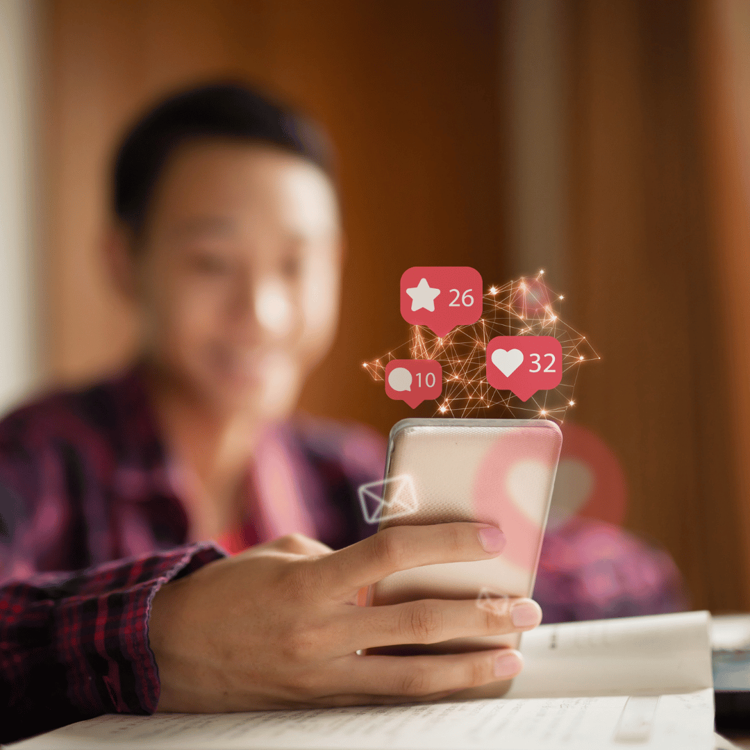 A person holding a smartphone with social media icons like likes, comments, and shares floating above it.