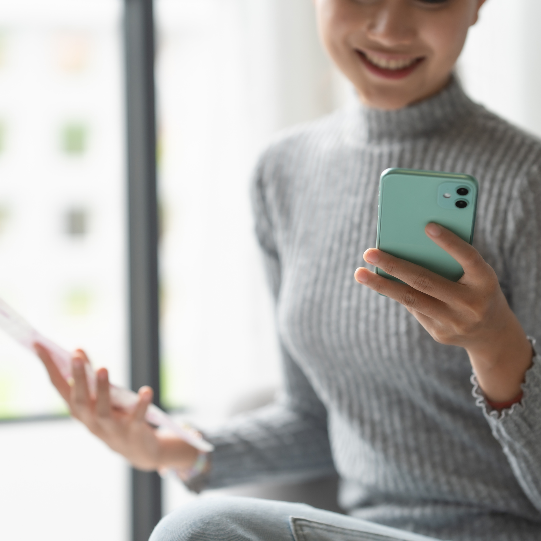A person in a grey turtleneck sweater holds a light green smartphone in their right hand and papers in their left hand.