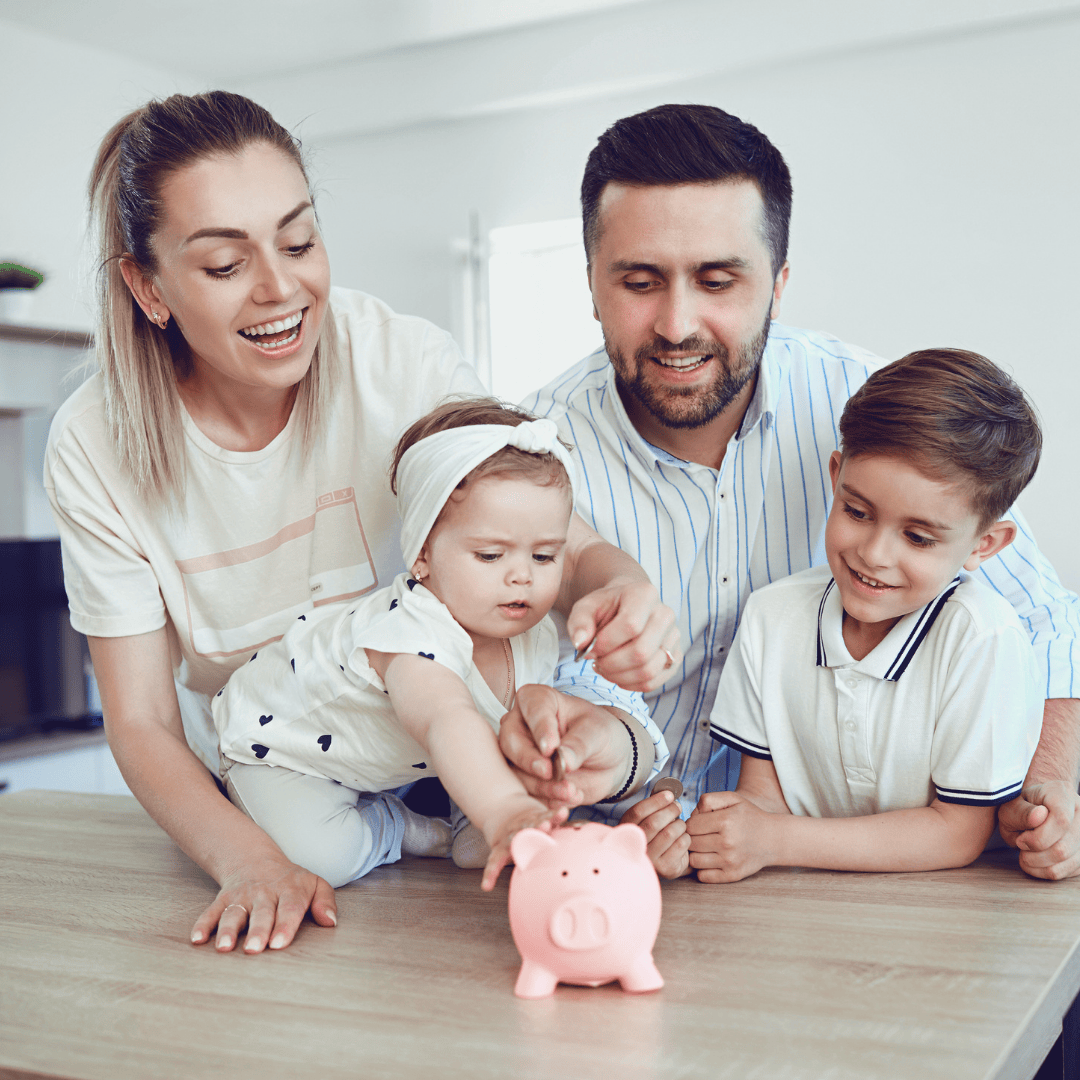 A family of four is gathered around a table, with the parents assisting their two children in putting coins into a pink piggy bank.