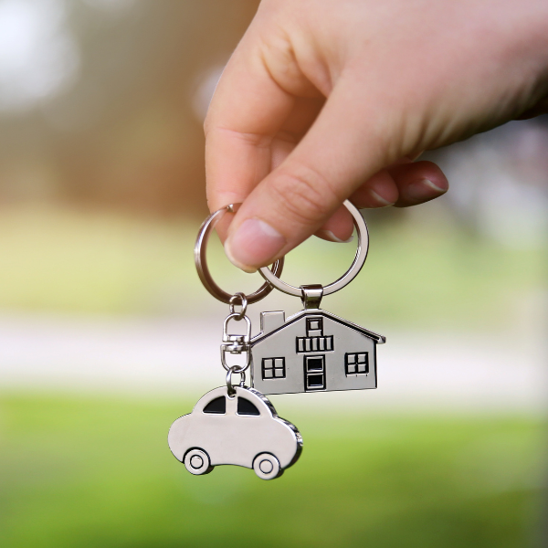 A hand holding two silver keychains. One is a car and the other is a house.